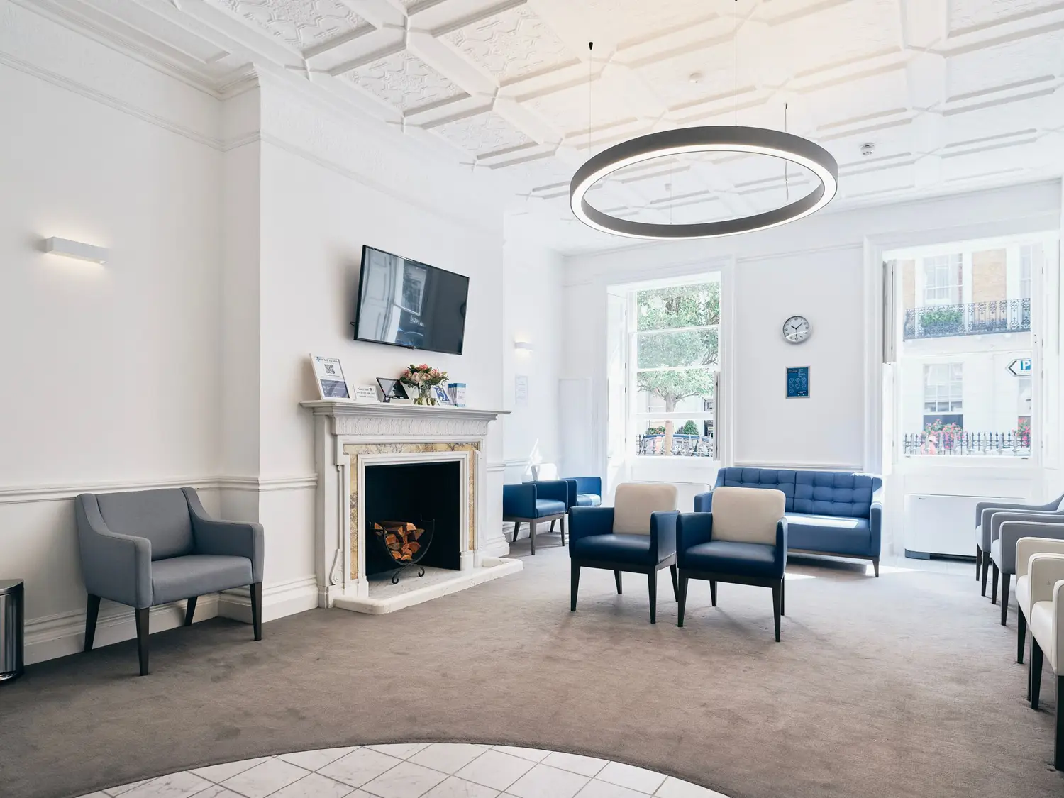 A modern waiting room in health assessment London, featuring gray and blue chairs, a fireplace, a wall-mounted TV, and large windows letting in natural light. The carpeted floor complements the decor of a clock and framed pictures, providing an inviting space for high-end health assessments.
