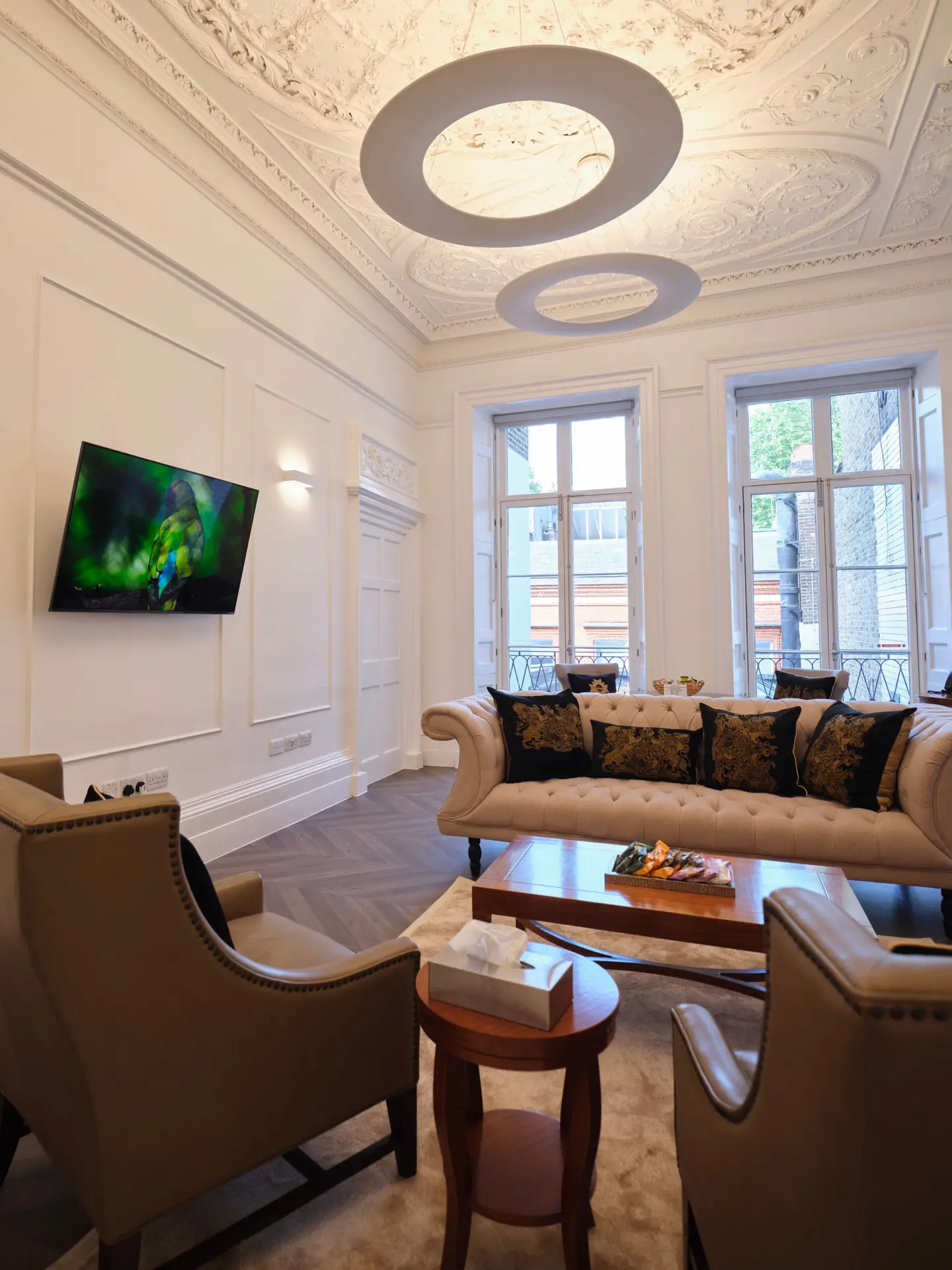 A luxurious living room with ornate ceilings features a sofa, armchairs, and a wooden coffee table. A wall-mounted TV displays a nature scene, perfect for relaxing after a health assessment in London. Large windows invite natural light, complementing the intricate moldings and floral-patterned cushions.