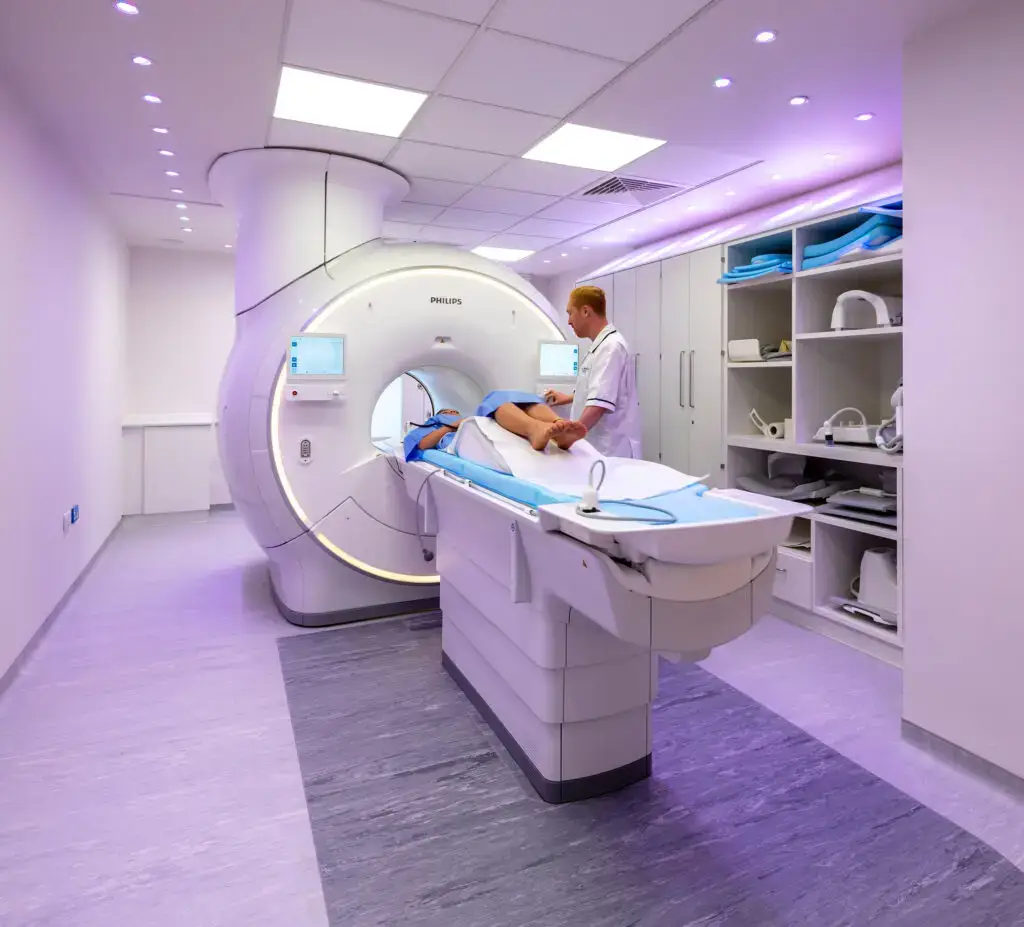 A technician operates an MRI machine in a brightly lit room, conducting a full body health scan. A patient lies partially inside the scanner. Shelves with medical equipment are visible on the right, while screens on the machine display vital information for this comprehensive checkup in London.