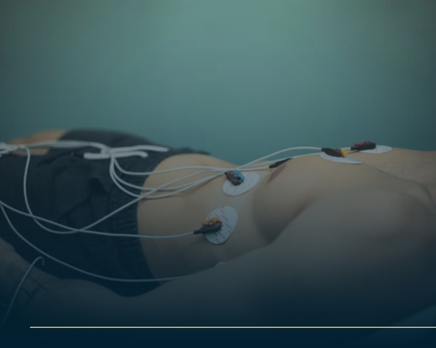 A person lies down for an ECG scan with electrodes attached to their chest and abdomen, all connected by wires. The soothing background is a soft teal, complimenting the patients dark shorts.