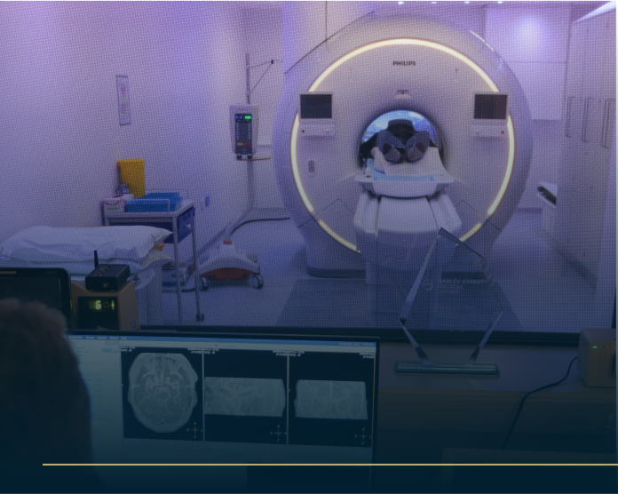 A person is inside an MRI machine in a medical room, surrounded by equipment and screens. A monitor displays images of brain scans. The room is well-lit with soft purple lighting.