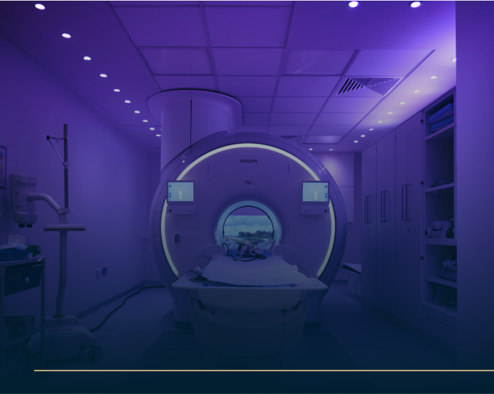 A modern MRI machine in a dimly lit room with a purple glow. The machines circular opening is illuminated, and there are screens on either side. Another room is visible through a window in the background.