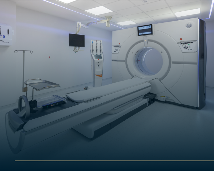 A modern medical imaging room featuring a CT scanner. The scanner has a circular opening with a sliding bed. Theres a monitor and control panel on the wall, and the room is brightly lit with white walls and flooring.