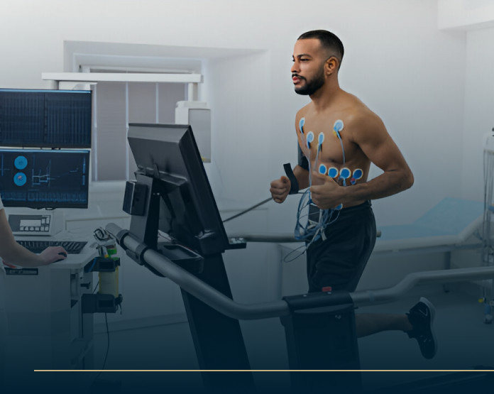 A man runs on a treadmill in a medical examination room, wearing electrode sensors on his chest. A technician monitors from a nearby computer displaying heart activity. The setting is bright and clinical.