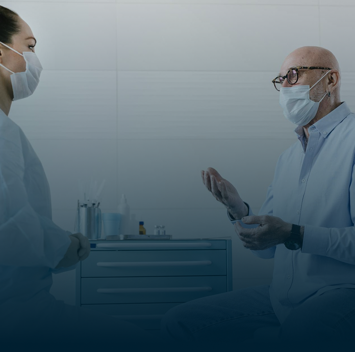 An older man wearing glasses and a mask talks to a female healthcare professional in a medical office. Both are seated and wearing masks, with medical supplies visible in the background.