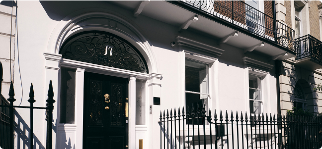 A white townhouse with a black ornate door featuring a gold knocker and letter slot, arched fanlight above, iron railings, and large windows, located on a sunny street.