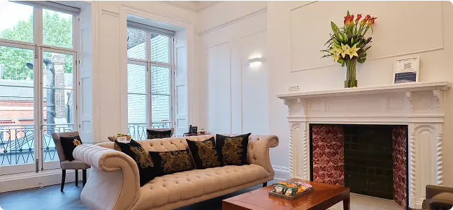 A bright, elegant living room with a cream sofa, black and gold cushions, a wooden coffee table, a white fireplace with a flower arrangement, and large windows letting in natural light.