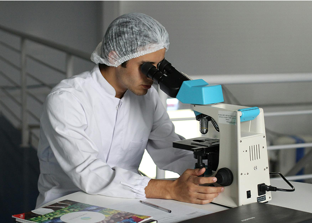 A person wearing a white lab coat and a hair net looks through a microscope in a laboratory setting, with documents and a pen on the desk nearby.