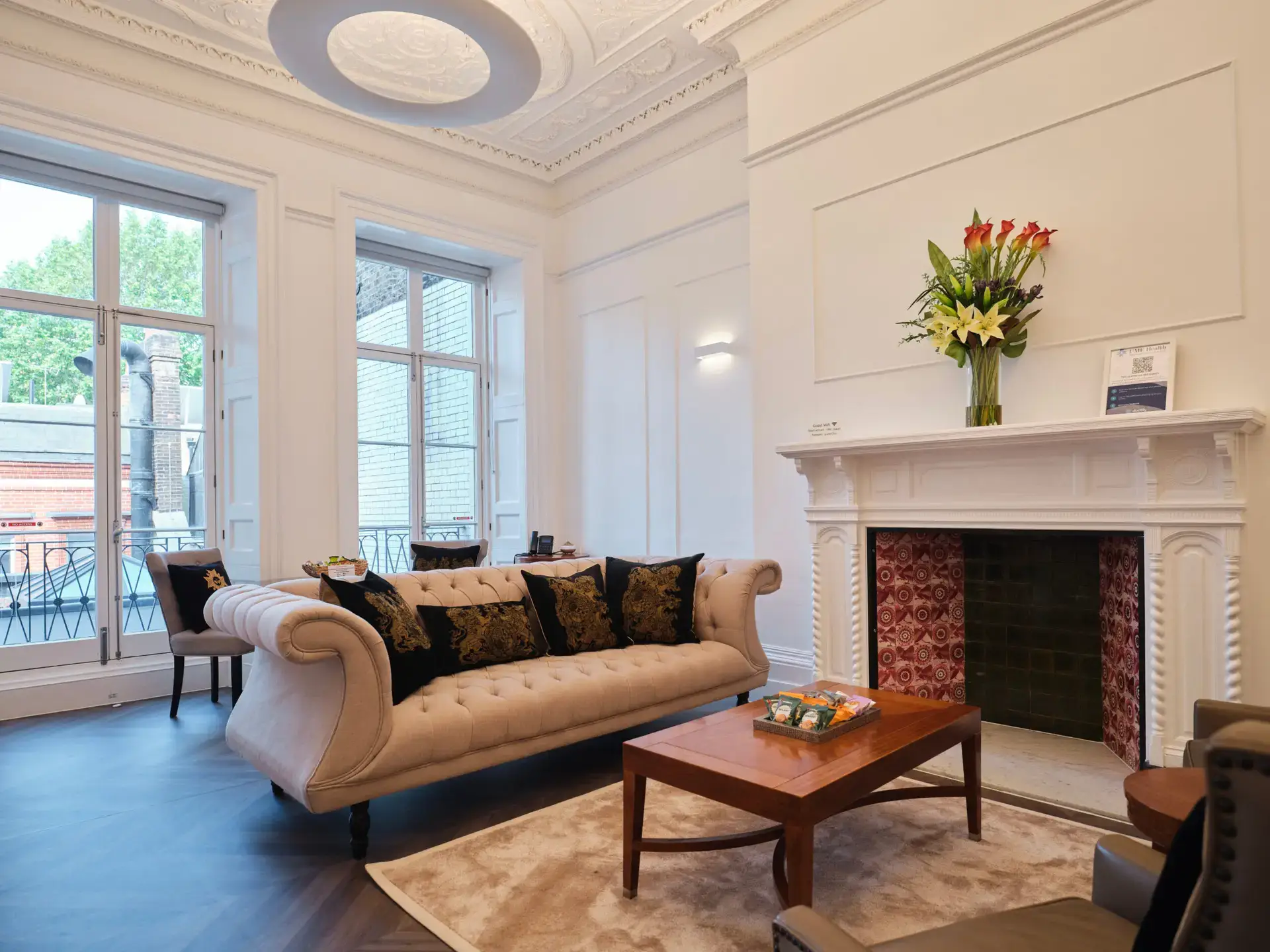 Elegant living room with a cream tufted sofa, black and gold pillows, wooden coffee table, and fireplace with red tile and floral arrangement. Large windows let in natural light and offer a view of the street outside.