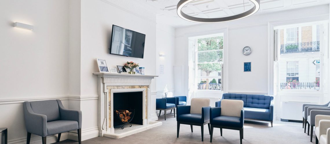 A modern waiting room in health assessment London, featuring gray and blue chairs, a fireplace, a wall-mounted TV, and large windows letting in natural light. The carpeted floor complements the decor of a clock and framed pictures, providing an inviting space for high-end health assessments.
