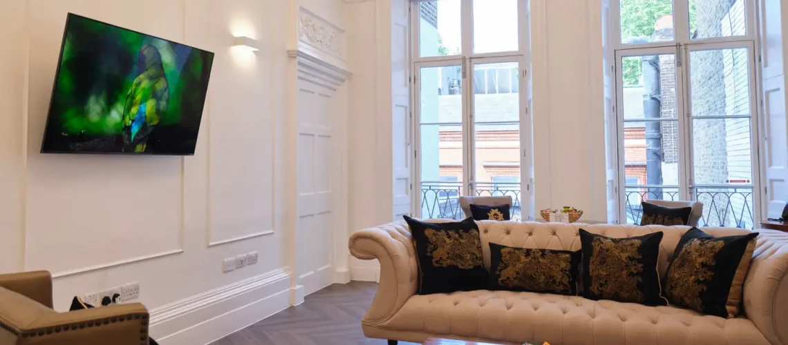 A luxurious living room with ornate ceilings features a sofa, armchairs, and a wooden coffee table. A wall-mounted TV displays a nature scene, perfect for relaxing after a health assessment in London. Large windows invite natural light, complementing the intricate moldings and floral-patterned cushions.