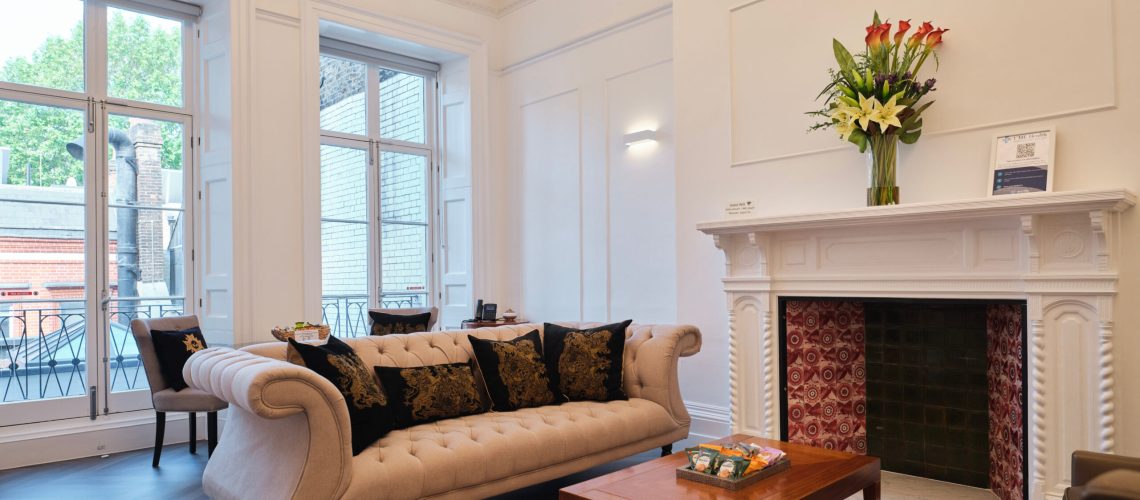 Elegant living room with a cream tufted sofa, black and gold pillows, wooden coffee table, and fireplace with red tile and floral arrangement. Large windows let in natural light and offer a view of the street outside.